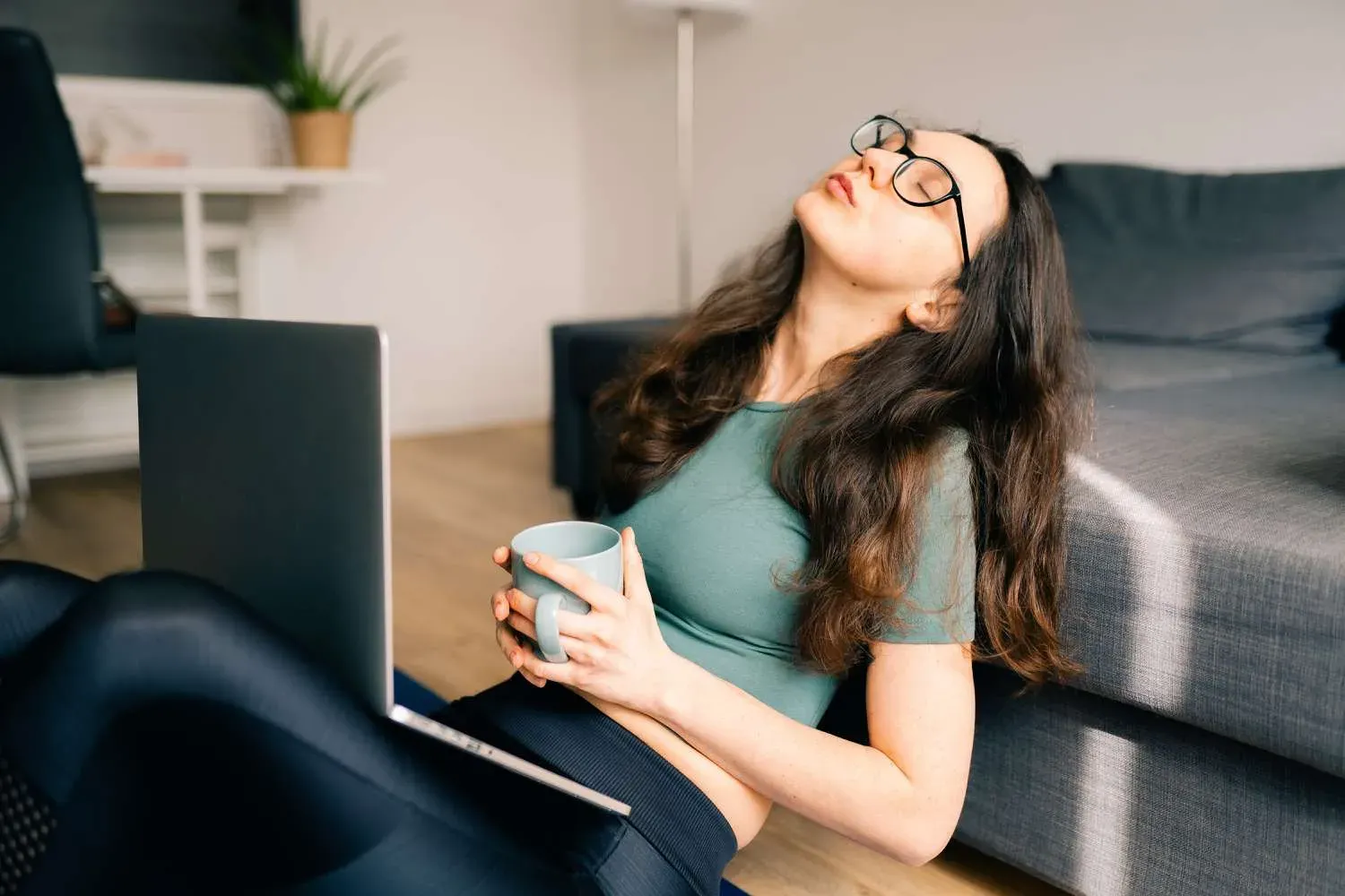 Mulher sentada no chão com o notebook no colo, cabeça pendendo para trás e olhos fechados de sono