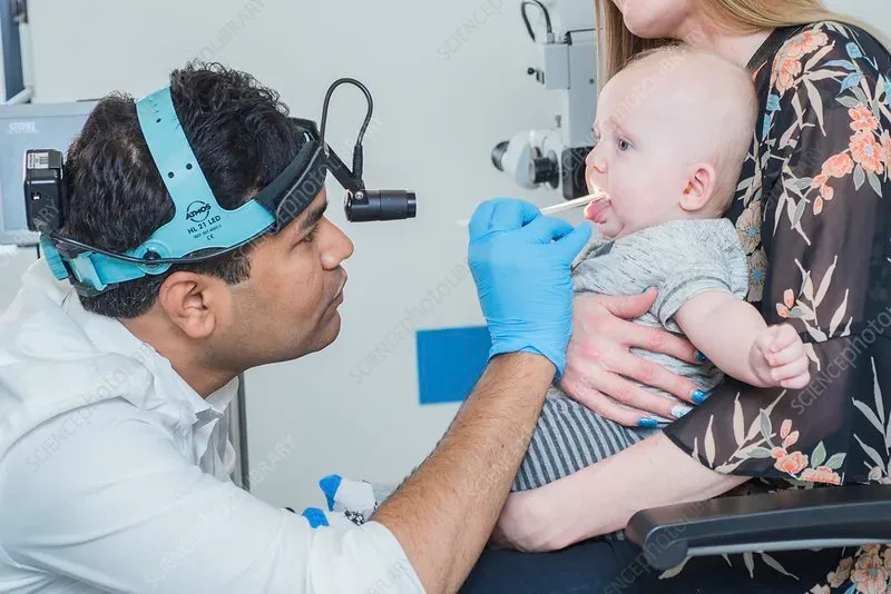 Otorrinolaringologista examinando a garganta de um bebê no colo da mãe, com lanterna de cabeça e abaixador de língua
